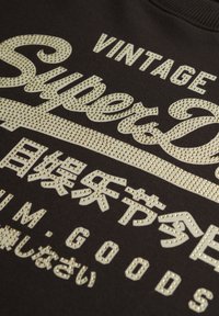 Zwarte sweatshirt met gouden glittergrafische tekst en accenten. Het ontwerp bevat een mix van Engelse en Japanse tekens. Gestructureerde afwerking.