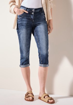Femme portant un pantalon capri en jean bleu délavé retroussé, une chemise beige à boutons, un top blanc et des sandales tissées avec des boucles.