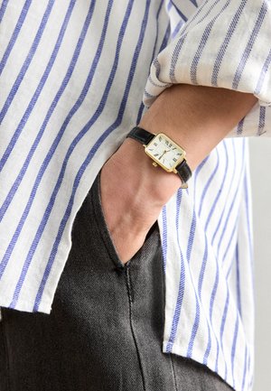 Reloj rectangular dorado con esfera blanca, correa de cuero negra, combinado con una camisa a rayas azul y blanca y pantalones gris oscuro.