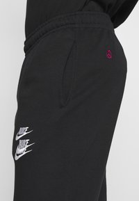 Svarta träningsbyxor med dragsko i midjan, med vita Nike-logotyper och en liten rosa fredssymbol broderad på sidan.