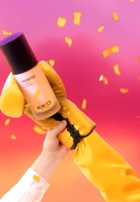 Mano sosteniendo un guante de boxeo amarillo que agarra una botella de KIKO MILANO Unlimited 24H Matte Foundation frente a un fondo rosa-naranja con confeti.