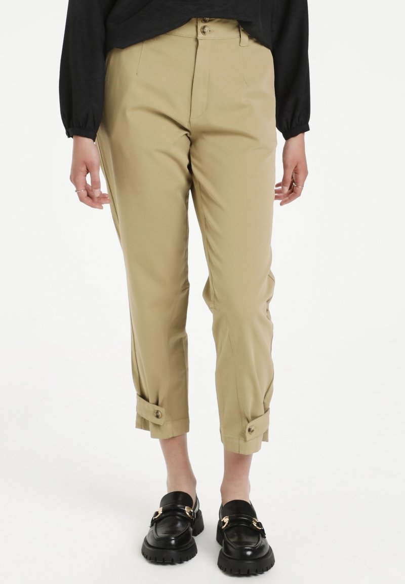 Beige beskurna byxor med en skräddarsydd passform, med uppvikta fållar som säkras med knappar. Bärs med svarta loafers. Strukturerad tyg.