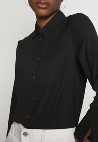 Camicia nera con bottoni realizzata in tessuto liscio, con colletto a punta, bottoni frontali e accenti a costine sulle maniche, abbinata a pantaloni di colore chiaro.