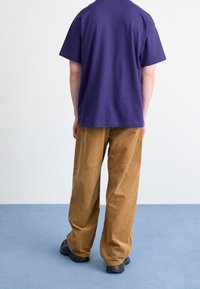 Lilla bomuldst-shirt og brune fløjlsbukser med løstsiddende pasform og horisontal ribstruktur, båret med sort fodtøj. Baggrunden er lysegrå.