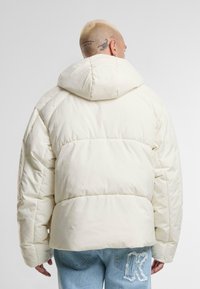 Jacket s kapuljačom, punjena s pjenom, u boji slonovače; sadrži kvadratni uzorak i ležeran kroj. Pogled straga otkriva teksturiranu tkaninu i šavove.