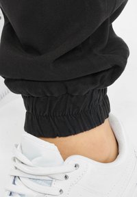 Gros plan sur une cheville portant un pantalon à poignets élastiques noirs et des baskets blanches lacées sur une surface claire.