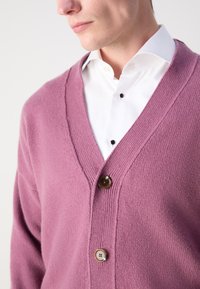 Cardigan s V-izrezom u mekom ružičastom pletenju, s teksturiranom površinom i dva smeđa gumba. Nosio se preko bijele košulje s crnim gumbima.