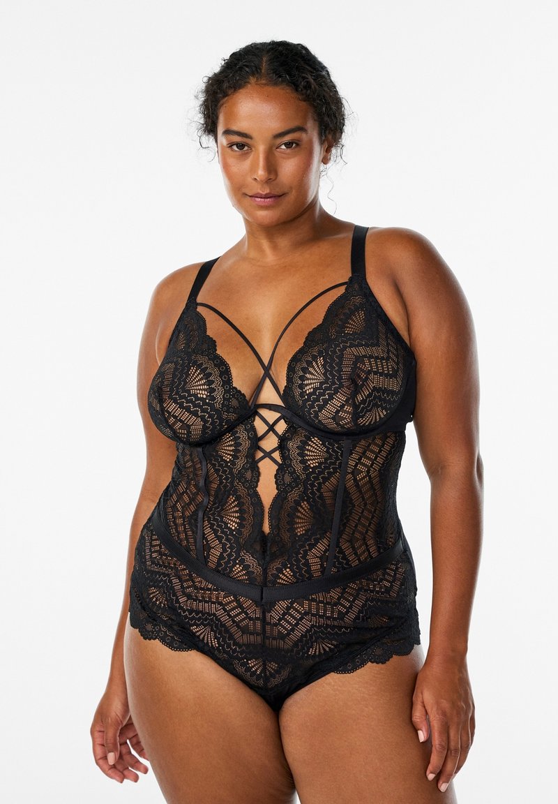 Femme grande taille portant un body en dentelle noire avec des bretelles croisées, debout sur fond blanc, regardant devant elle avec confiance.