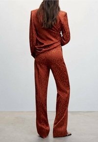 Femme aux longs cheveux bruns vue de dos, portant un costume en satin à motifs couleur rouille, composé d'un pantalon à jambes larges et d'un blazer.