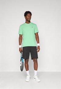 Zelené bavlnené tričko s okrúhlym výstrihom, skombinované s čiernymi šortkami. Model drží tenisovú raketu a má obuté biele ponožky a topánky.