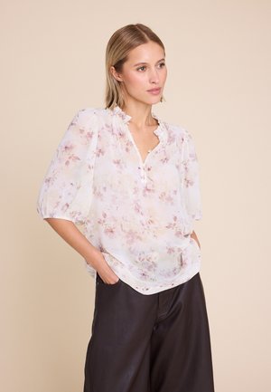 Kvinne i gjennomsiktig hvit bluse med rosa blomster og puffermer, iført mørke bukser med vide ben, stående med hendene i lommene.