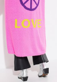 Helle pinke Strickjacke mit "LOVE" in Gelb und einem Friedenszeichen, getragen über schwarzen Kunstlederhosen und silbernen Absatzstiefeln.