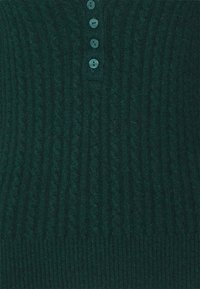Jersey de punto verde oscuro con un patrón de cable texturizado, cinco pequeños botones en el cuello y un dobladillo acanalado.