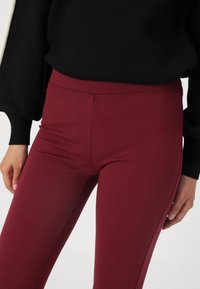 Leggings bordeaux en tissu extensible, dotés d'une taille haute avec une texture lisse et des coutures minimales sur les côtés. Haut noir partiellement visible.