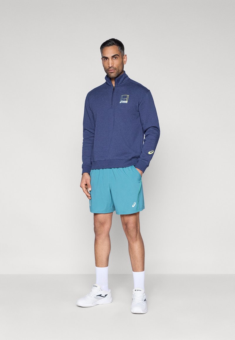 Pull à fermeture éclair bleu marine avec un logo, associé à un short bleu clair. Le mannequin porte des baskets blanches et des chaussettes basses, debout sur un fond neutre.