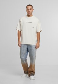 Vyras stovi vilkėdamas per didelį off-white spalvos marškinėlį su užrašu "2Y STUDIOS" ir išblukusiomis dviem tonais plačiomis kelnėmis bei baltomis sportbačiais.