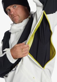 Hvid og sort jakke med gule accenter, der har en lynlåset mesh indvendig lomme. Modellen trækker stoffet tilbage for at vise indersiden.