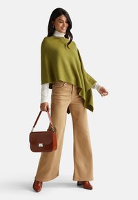 Poncho verde oliva, pantaloni beige a gamba larga e tacchi alti marroni a punta. Borsa marrone con motivo texturizzato e chiusura in argento. Dolcevita a strati.