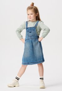 Denim-Latzkleid in Hellblau mit einer Tasche, getragen über einem mintgrünen, gestreiften Langarmoberteil. Weiße Sneakers mit schwarzen Streifen auf den Socken.