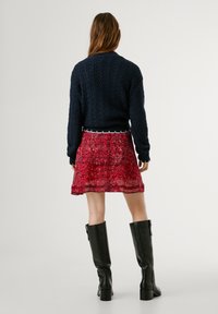 Jūras spēku kabelī adīts džemperis ar ribotu apakšmalu, sakombinēts ar slāņotu sarkanu ziedu svārku. Melni ceļgalu auguma zābaki ar blokveida papēdi papildina apģērbu.