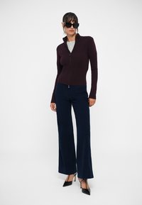 Burgundijas krāsas zip-up džemperis ar augstu apkakli, apvienots ar jūras zila krāsas platām biksēm un melniem norādītas pirkstgalu apaviem; gluda auduma un pieguļoša dizaina.