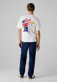 Witte katoenen t-shirt met verschillende kleurrijke merkl logo's op de achterkant, gecombineerd met blauwe spijkerbroek en witte schoenen.