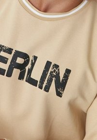 Camisa de cuello redondo beige con un gran texto negro desgastado que muestra parcialmente la palabra "BERLÍN" en el pecho.