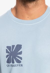 Detailní záběr na světle modré tričko Quiksilver, které nosí muž. Tričko má na hrudi tmavě modré abstraktní logo a název značky.