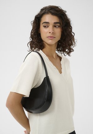 Ženska s kodrastimi lasmi, oblečena v kremni pleteni pulover, nosi črno ramensko torbico v obliki polmeseca, gleda vstran pred enobarvnim ozadjem.