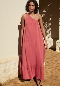 Abito maxi rosa a spalla singola con sottile spallina e dettaglio arricciato. Tessuto fluente e orlo a strati. Indossato con sandali neri, su uno sfondo di parete testurizzata.