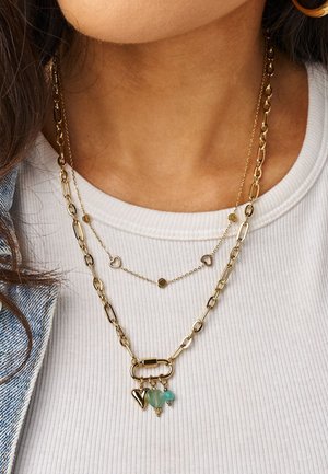 Zwei goldene Halsketten in Lagen an einer Frau, die ein weißes Rippenoberteil und eine Jeansjacke trägt; eine Halskette hat Herzanhänger und grüne Anhänger.