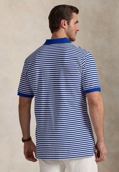 Polokošile z modro-bílého pruhovaného materiálu. Obsahuje modrý límec a krátké rukávy s mírně delším zadním dílem a bočními rozparky.