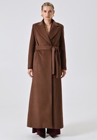 Bruine, lange wollen jas met een riem, zijzakken, piekflappen en een soepele textuur. Draagt een bordeauxkleurige top en zwarte puntige schoenen.