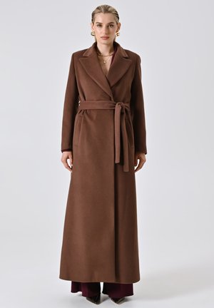 Cappotto lungo marrone in lana con cintura, tasche laterali, rever a punta e una texture liscia. Indossato sopra un top bordeaux e scarpe nere a punta.