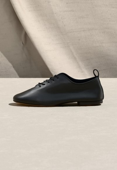 Black leather lace-up flat shoe with rounded toe and pull tab on back, placed on light surface against a textured beige fabric background.