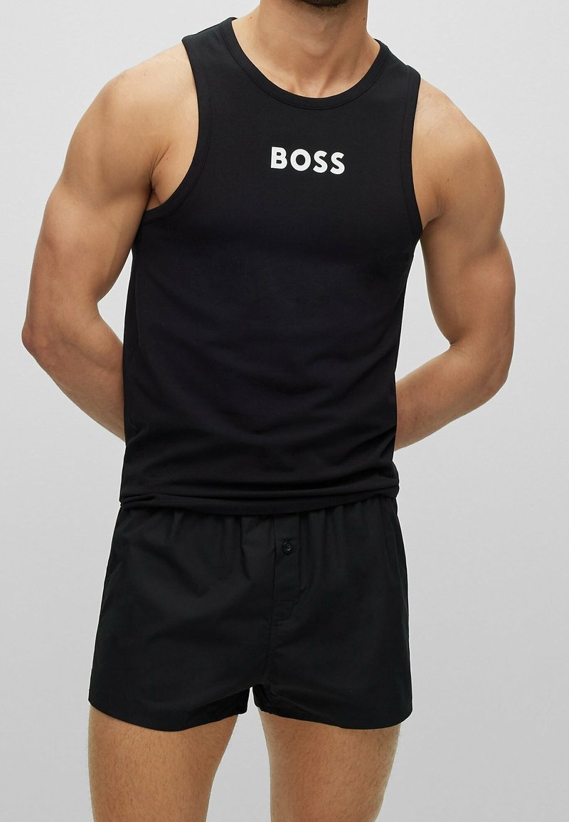 En muskulös man står med händerna bakom ryggen iklädd en svart linne med texten "BOSS" och svarta boxershorts mot en enfärgad bakgrund.