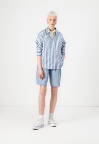 Striped blue and white button-up shirt with long sleeves, paired with light blue denim shorts and white high-top sneakers.