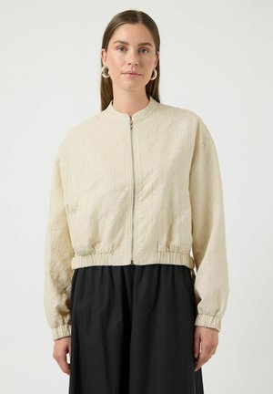 Młoda kobieta z prostymi włosami ubrana w beżową, pikowaną kurtkę na suwak i czarną spódnicę, stojąca na tle jednolitego jasnego tła.