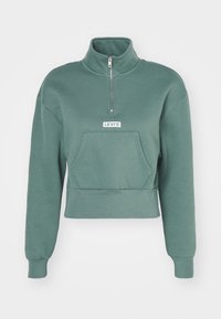 Felpa verde cropped con colletto alto, mezza zip, tasca frontale e patch del logo Levi's bianco. Tessuto morbido e vestibilità rilassata.