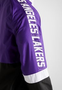 Lila vindjacka med en svart nederdel, vit kant och stora vita bokstäver som säger "Los Angeles Lakers" längs ärmen.