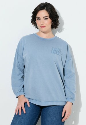 Ung kvinde med kort krøllet hår iført en lyseblå langærmet skjorte med teksten "LEO LOVE" og mørkeblå jeans, stående mod en hvid væg.