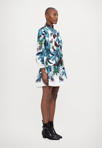 Blumendruckkleid in Weiß, mit blauen und grünen Blättern, langen Ärmeln, tailliertem Schnitt und fransigem Saum. Kombiniert mit schwarzen Stiefeletten.