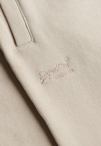 Beige Baumwollstoff mit glatter Textur, der ein gesticktes "SuperDry"-Logo in einem dezenten Farbton zeigt. Zwei vertikale Paneeldetails sind vorhanden.