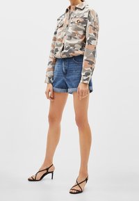 Camicia button-up camouflage nei toni beige, grigio e arancione; abbinata a pantaloni corti in denim e sandali neri con cinturini. Vestibilità casual e design rilassato.