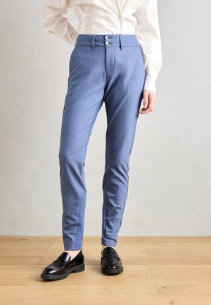 Mujer con pantalones azules de corte ajustado y doble botón, y mocasines negros, de pie sobre un suelo de madera frente a una pared clara.