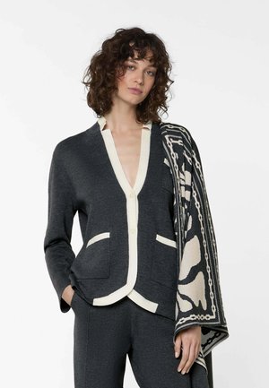 Vrouw met krullend haar draagt een donkergrijze gebreide cardigan met witte biezen, bijpassende broek en legt een zwart-witte sjaal met patroon over haar schouder.