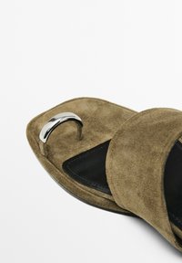 Semišové sandály na klínku v olivově zelené barvě s černým vnitřním podšitím, se stříbrným metalickým akcentem na pásku. Plochý design s otevřenou špičkou.