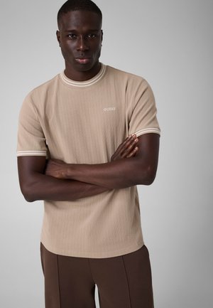 Jeune homme à la peau foncée portant une chemise beige à manches courtes avec bordure blanche et un pantalon marron, debout les bras croisés sur fond gris.