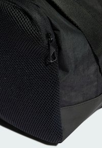 Bolsa negra con un panel lateral de malla texturizada, cierre de cremallera y una correa resistente. El material es suave con un acabado mate.