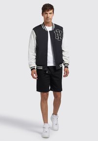 Zwart-wit varsityjack met een letterembleem, lange mouwen, geribde manchetten en een drukknoopsluiting. Combinatie met zwarte shorts en witte sneakers.